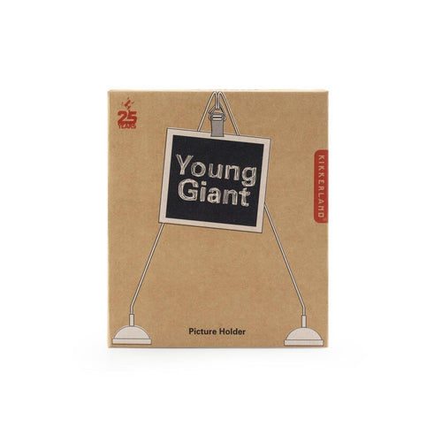 Brown box with illustration of a photo being held by a clip. Clip is at the top where two stands meet in a triangle shaped point. Illustrated photo reads 'young giant'. Kikkerland logo sits to right of the packaging. '25 years' is written in red in the top left. 'Picture holder' is written in black across the bottom. 