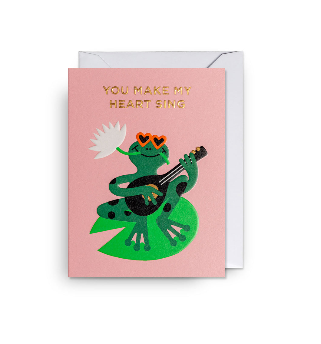 Pink card, gold lettering. Illustration of a frog wearing heart sunglasses, holding a banjo, sitting on a lily pad.