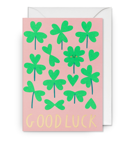 Light pink card with green clovers and a smiling four leaf clover. 'Good luck' is written in gold letters'. card sits on white envelope. 