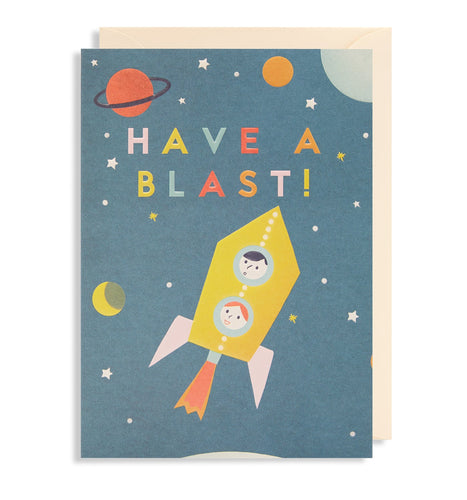 Blue card with white envelope inside. Illustration is of two light-skinned childrens faces (one with black hair, one with ginger hair) inside a yellow rocket. There are little stars, and planets around the rocket. In colourful capital letters 'have a blast!' is written across the top.