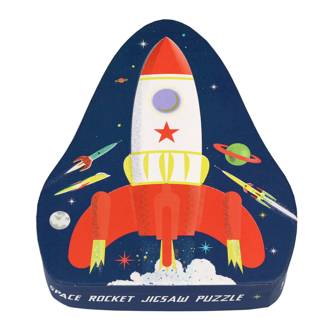 Puzzle box is a curved triangle shape. Box is dark blue with illustrated white rocket with red and yellow accents. Fire and smoke comes from the bottom of the rocket. In the background are two colourful ringed planets, the moon, stars, and three other colourful rockets. Along the rim of the lid reads 