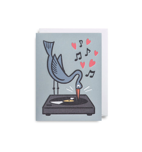 Dusty blue card with white envelope tucked inside. An illlustration of a tall blue bird leaning over and using it's beak as a needle on a record player. Music notes and pink hearts float upwards. 