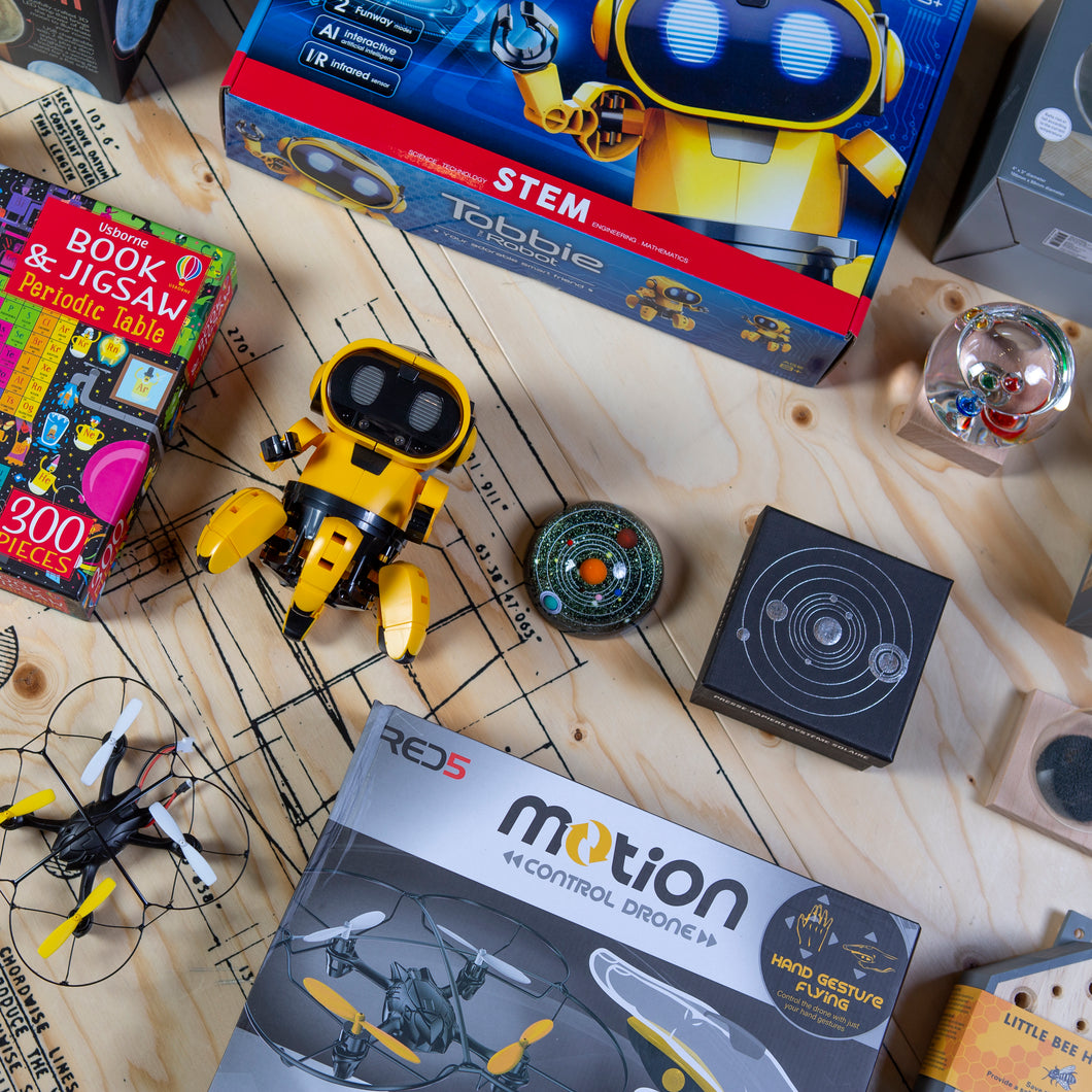 Shop items on a wooden surface. Products seen include Tobbie Robot box and Robot lying flat on the surface, motion control drone with drone out of the box, periodic table jigsaw puzzle, planetary paperweight and box, galileo thermometre and box. More products can be seen continuing out of the image. 