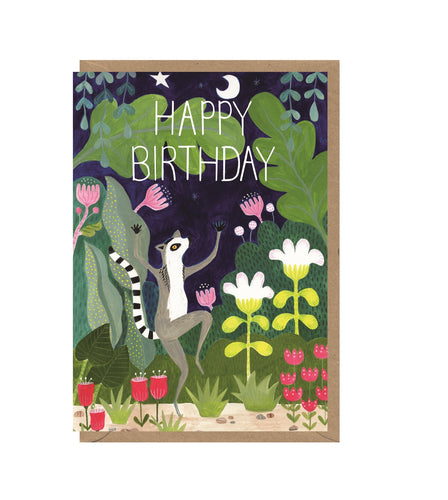 Card with a brown kraft envelope tucked inside. Illustration is of a dancing ring-tailed lemur surrounded by plants and a dark night sky with moon and stars. Card reads 'happy birthday' in capital white letters.