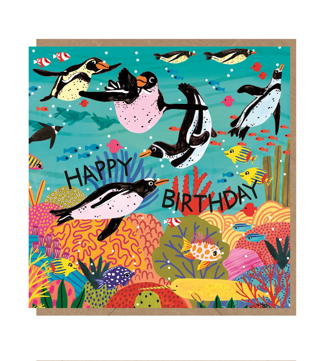 Square card with brown kraft envelope tucked inside. Illustration on the card is of 8 penguins swimming in a coral reef with different types of fish. The words 'happy birthday' are curved in the middle of the card with capital black letters.