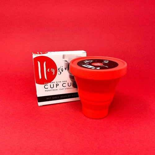 Red tapered pot with a lid in front of white packaging. Packaging reads 'Hey Girls, buy one give one, cup cup, menstrual cup steriliser.' 
