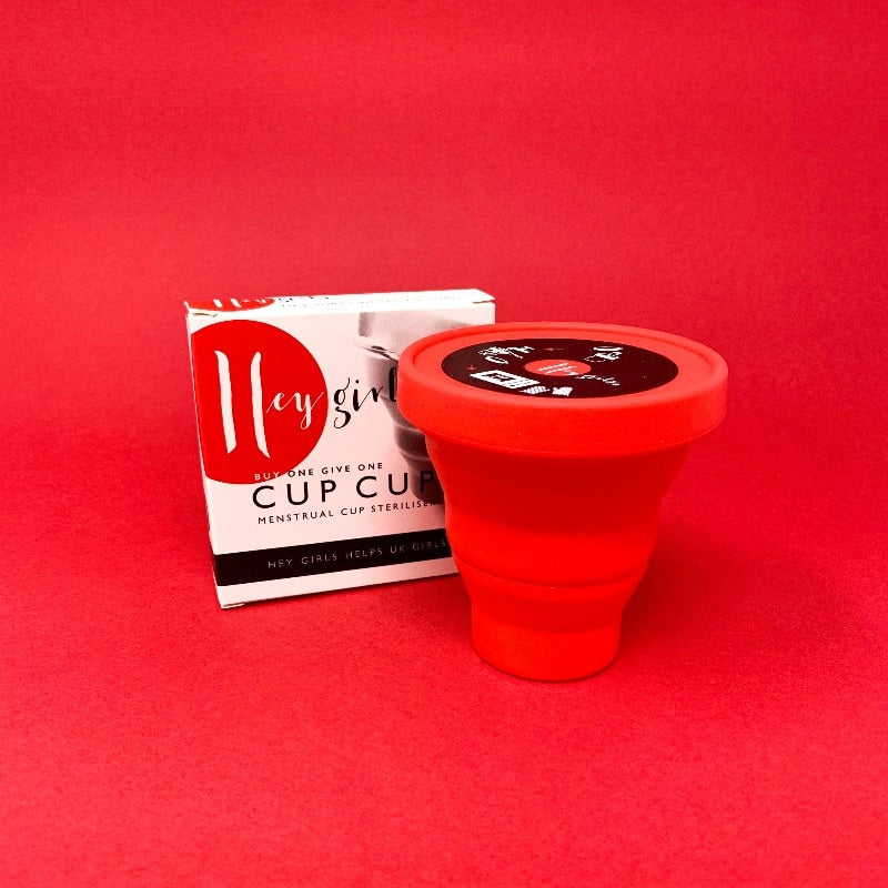 Red tapered pot with a lid in front of white packaging. Packaging reads 'Hey Girls, buy one give one, cup cup, menstrual cup steriliser.' 