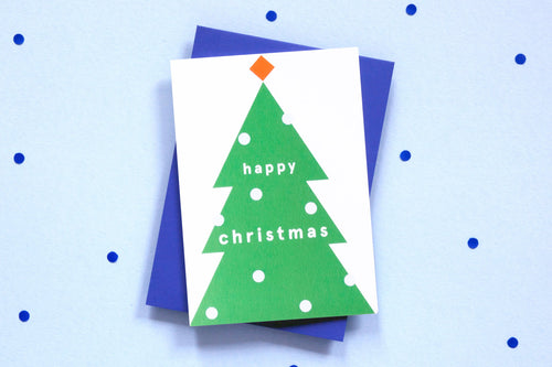 Card on top of blue envelope. White card with green Christmas tree. Baubles on the tree are white circles. Lettering on the tree reads 'happy christmas' in lower case letters. On top of the tree is an orange diamond.