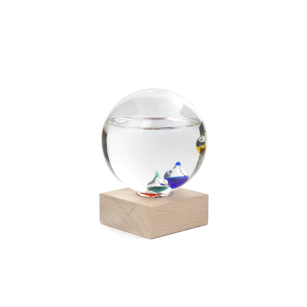 Glass Galileo Thermometer is a globe rests on square wooden block. Smaller globes inside are multi-coloured (yellow, blue, teal, red can be seen) and floating at different heights. Liquid in the globe is three quarters full.