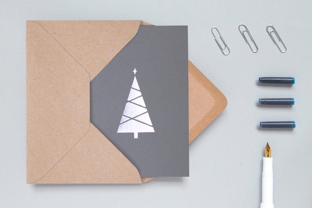 Dark grey card with silver foiled Christmas tree. Card is partially inserted into brown envelope. Foil tree is crossed by grey lines to indicate lights or garlands. On top of the tree is a 4 pointed star in the same silver foil. 