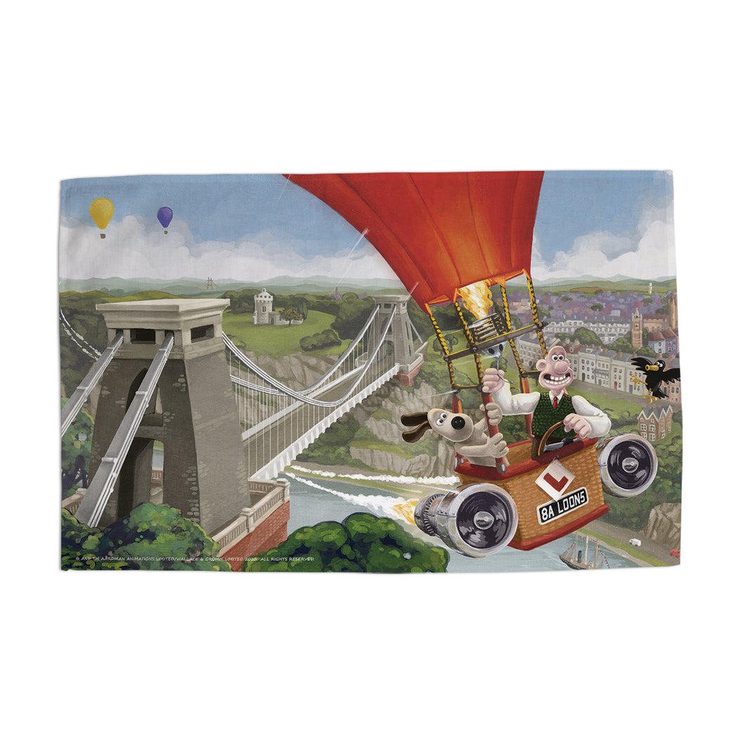 This Aardman tea towel features Wallace (a white man) & Gromit (a dog) flying over the Clifton Suspension Bridge in a hot air balloon. 