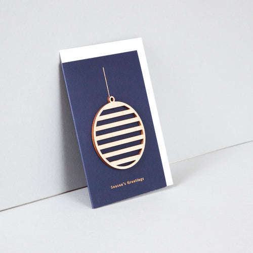 Blue portrait card with wooden ornament attached and white envelope inside. Card reads 'Season's Greetings' across the bottom in small gold letters. Ornament is attached and has a string for hanging. Ornament is round and striped. 
