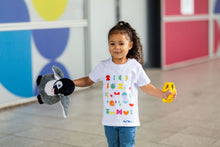 Load image into Gallery viewer, Child smiles off camera with toys in her hands. She is wearing a white t-shirt with colourful shapes and blue jeans. 
