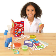 Load image into Gallery viewer, A medium-skinned girl in a lab coat holds the Science Lab Book in one hand and a rainbow bouncy ball in the other. The contents of the box are laid out on the wooden surface in front of her. These include, paper, goggles, test tubes, petri dish and pot of something green.

