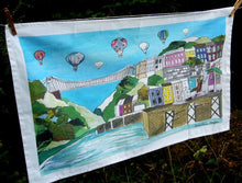 Load image into Gallery viewer, The unfolded tea towel has a white border around a colour illustration of the Clifton Suspension Bridge and surrounding colourful houses with the river below. Hot air balloons fill the sky.
