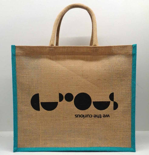 Brown bag with brown handle and teal border. In the centre of the bag is the black We The Curious logo, with we the curious written upside down, and the word 'curious' spelled out in circles and half circles. 