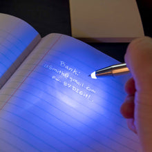 Load image into Gallery viewer, A white hand holds the invisible pen over a page, it&#39;s lit up to see some writing.
