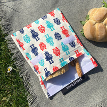 Load image into Gallery viewer, Robot baggie is open with bread and breadsticks inside. Two velro strips can be seen on the bag and flap. Bag sits on a blanket on some grass with a sandwich beside. 
