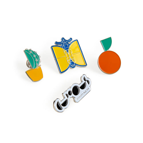 4 enamel pin badges. Designs include black and white We The Curious logo, blue and yellow satellite, green cactus in yellow pot, and orange mandarin with green leaf.