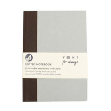 Load image into Gallery viewer, Notebook is grey with brown spine. Notebook is wrapped with paper band packaging. Packaging shows &quot;Vent for Change&quot; logo, and reads &quot;dotted notebook. sustainable stationery with style. Notebook made from recycled board and 100% sustainable paper.
